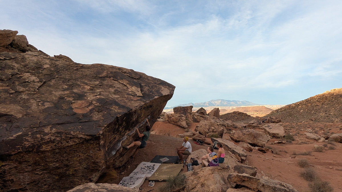A scenic desert landscape with large boulders, reddish-brown sandy ground, distant mountains, and a blue sky with wispy clouds. A few small people are visible climbing and sitting near the rocks in the middle distance.